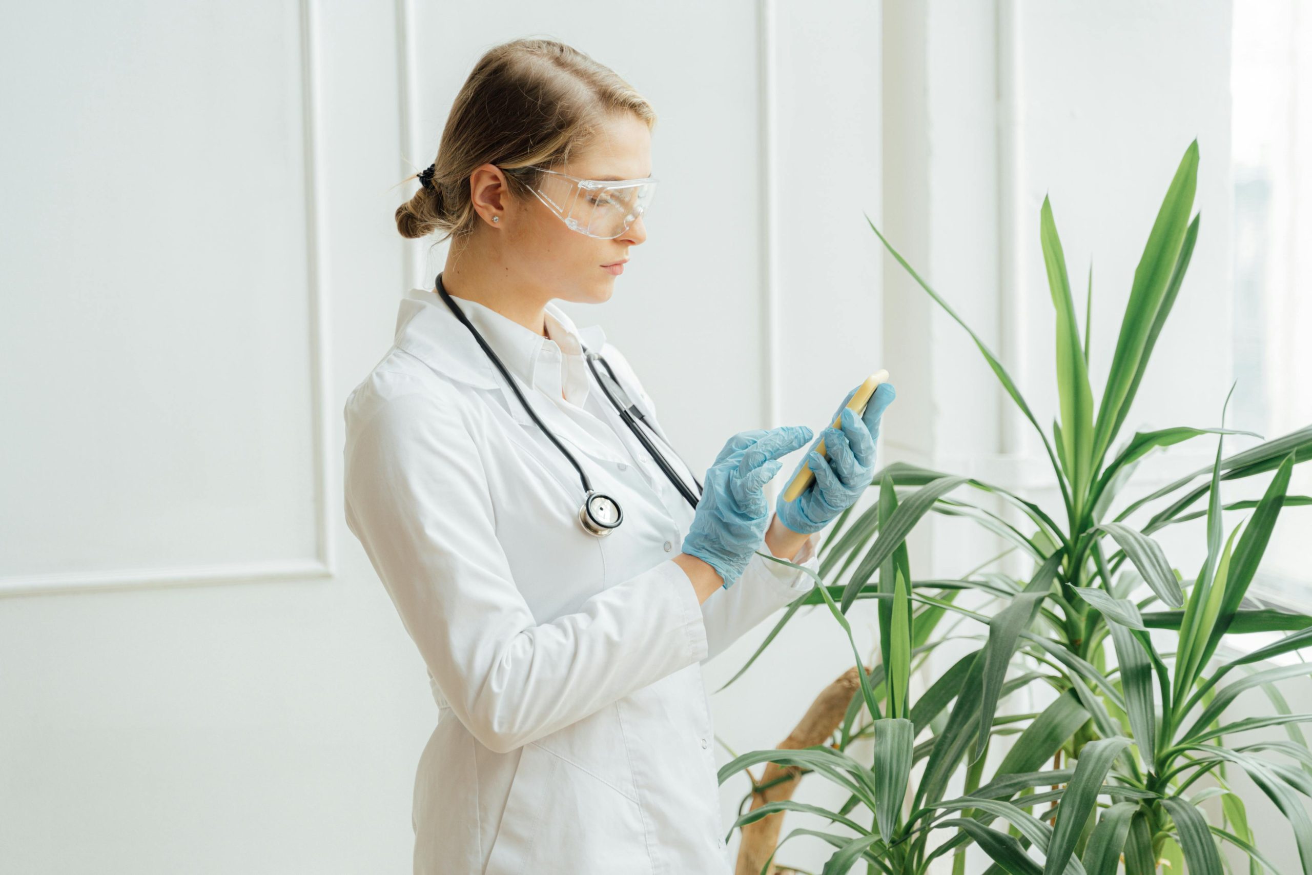 A female doctor in protective gear using a smartphone indoors beside green plants.
